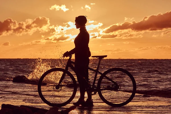 Silhouette di una ragazza con una bicicletta su una costa di mare su uno sfondo di tramonto . — Foto Stock