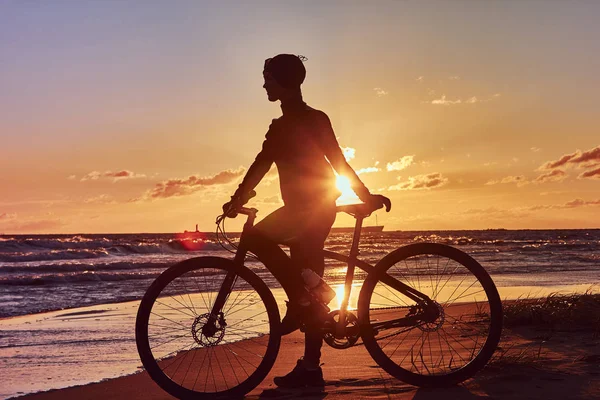 彼女は自転車の側に立って、海の海岸に夕日を楽しんで女性サイクリスト. — ストック写真