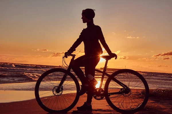 彼女は自転車の側に立って、海の海岸に夕日を楽しんで女性サイクリスト. — ストック写真