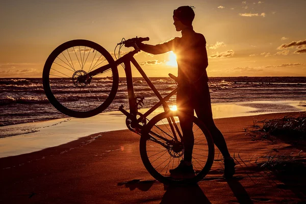 Silhouette of a woman holds a bicycle on a sea coast on a sunset background. — Stock Photo, Image