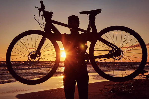 Silhueta de uma mulher segura uma bicicleta em uma costa do mar em um fundo por do sol . — Fotografia de Stock