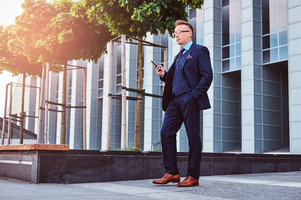 Retrato de un hombre de negocios con estilo y confianza vestido con un traje elegante sostiene un teléfono inteligente y mirando hacia otro lado mientras está parado al aire libre contra un fondo de rascacielos . —  Fotos de Stock