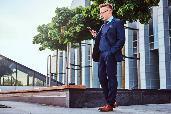 Porträtt av en säker eleganta affärsman klädd i en elegant kostym med en smartphone samtidigt som står utomhus mot skyskrapa bakgrund. — Stockfoto