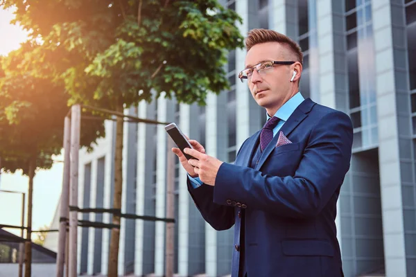 Portret pewnie biznesmen stylowe, ubrany w elegancki garnitur posiada smartfona i szukasz drogi na zewnątrz stojąc na tle drapacz chmur. — Zdjęcie stockowe