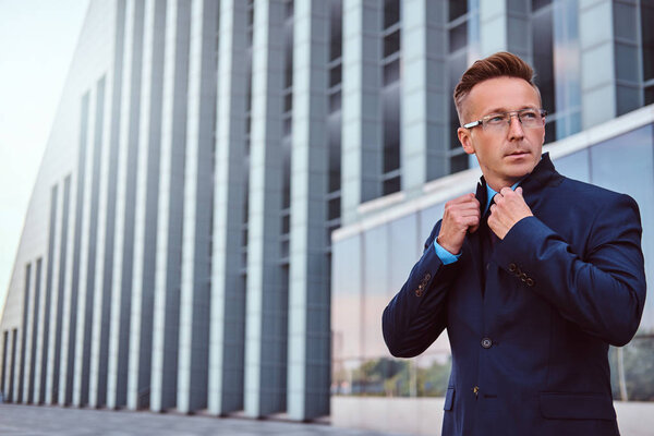 Portrait of a confident man in elegant suit and glasses correct his jacket while standing against skyscraper background.