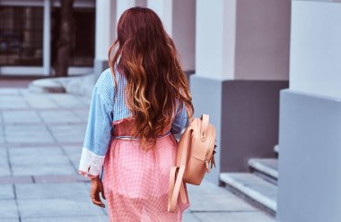 Arkadan görünüm trendy kıyafetler giymiş uzun kahverengi saçlı güzel bir kadının sokakta duran bir sırt çantası tutar.