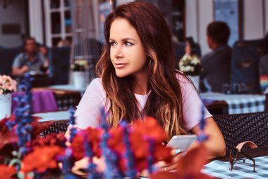 Uzun kahverengi saçlı orta yaş işkadını portresi açık bir kafede otururken bir akıllı telefon tutar.