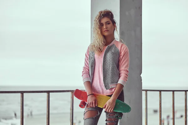 迷人的年轻女孩 金发女郎身着粉红色夹克 持有一个滑板附近的护栏 反对黑海和白色海岸背景 — 图库照片
