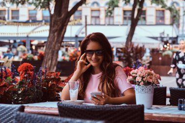 Gülümseyen bir orta yaş iş kadını açık kafede otururken bir smartphone kullanarak güneş gözlüğü uzun kahverengi saçlı portresi.