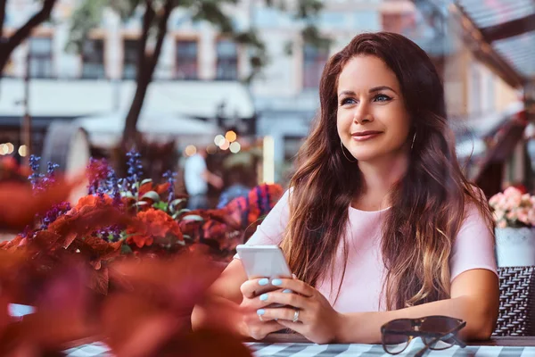 Happy middle age businesswoman with long brown hair holds a smartphone while sitting at an outdoor cafe.