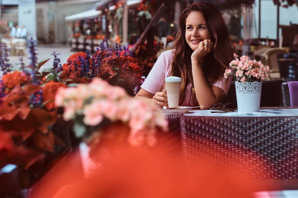 Happy middle age businesswoman with long brown hair wearing a pink dress holds glass of cappuccino while sitting at the outdoor cafe.