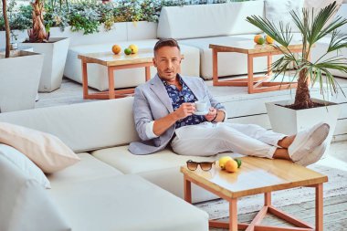 Bir Divan şehir wharf arka planı açık kafede otururken bir fincan kahve ile modern şık giysiler giymiş şık saç moda başarılı adam tutar.