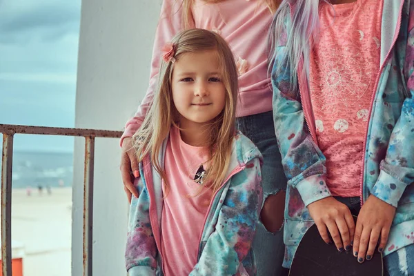 Portréja egy aranyos kislány, szőke haj öltözött a rózsaszín pólót és kék kapucnis álló családjával a korláthoz ellen tengerpart közelében. — Stock Fotó