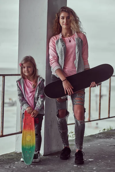 Hermosa chica con el pelo rubio y su hermana pequeña vestida con ropa de moda sostiene monopatines posando cerca de una barandilla contra una costa del mar . — Foto de Stock