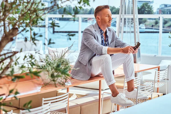 成功时尚男士穿着现代高雅的衣服坐在户外咖啡馆的桌子上, 在城市码头的背景下. — 图库照片