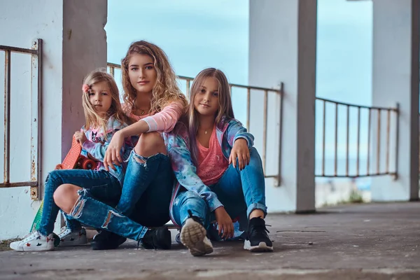 Hermosa chica con el pelo rubio y sus hermanas pequeñas vestidas con ropa de moda sentadas juntas en patinetas posando cerca de una barandilla contra una costa del mar . — Foto de Stock