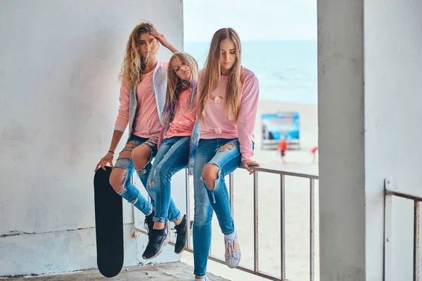 Portret van drie zusters gekleed in trendy kleding zit op een leuning tegen een Zeekust. — Stockfoto