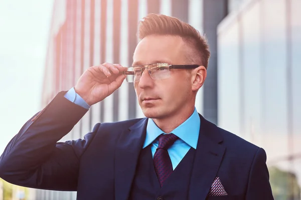 Retrato de cerca de un hombre de negocios con estilo y confianza vestido con un traje elegante mirando hacia otro lado y corregir sus gafas mientras está parado al aire libre contra un fondo de rascacielos . —  Fotos de Stock