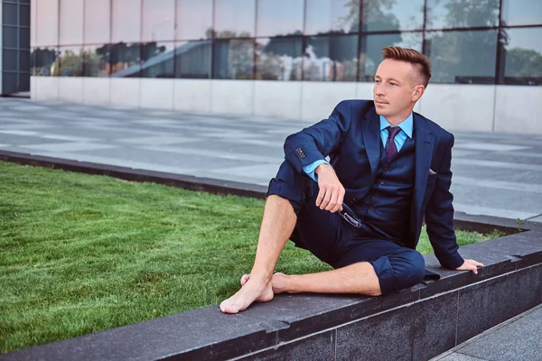 Empresário confiante vestido em um terno elegante segura seus óculos enquanto sentado descalço contra o fundo da paisagem urbana . — Fotografia de Stock