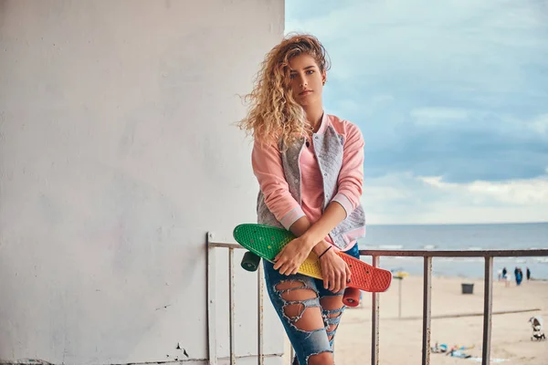 有魅力的年轻女孩 金发女郎身穿粉红色夹克 手持滑板 靠近海边的护栏 — 图库照片