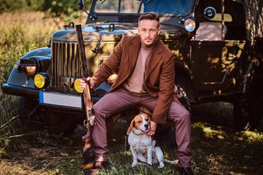 Şık giysileriyle avcı av tüfeği tutan ve retro bir askeri araç ormandaki yaslanmış iken onun beagle köpek ile birlikte oturur.