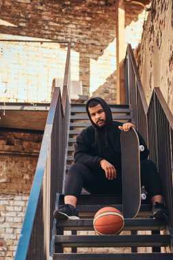 Bir siyah hoodie kılığında sakallı hippi adam koyu tenli ve spor şort bir Getto merdivenlerde otururken bir kaykay tutar.