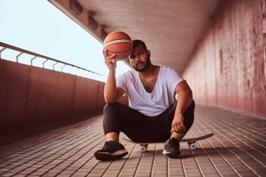 Dalgın bir esmer adam portresi beyaz bir gömlek giymiş ve spor şort tutan bir basketbol footway köprü altında bir kaykay üzerinde otururken.