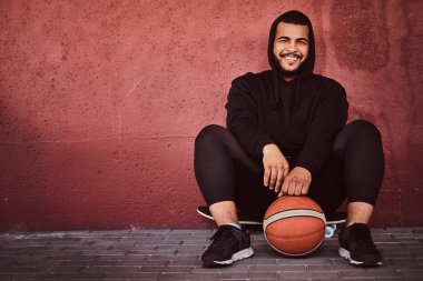 Neşeli Afrikalı-Amerikalı basketbol ile bir kaykay üzerinde oturan ve duvara yaslanmış bir siyah hoodie ve spor şort giymiş adam sakallı.