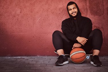 Neşeli Afrikalı-Amerikalı basketbol ile bir kaykay üzerinde oturan ve duvara yaslanmış bir siyah hoodie ve spor şort giymiş adam sakallı.