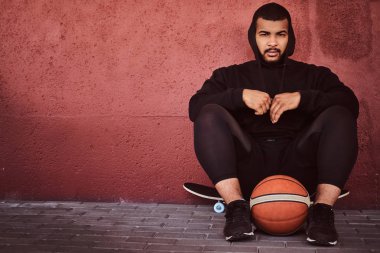 Afrikalı-Amerikalı basketbol ile bir kaykay üzerinde oturan ve duvara yaslanmış bir siyah hoodie ve spor şort giymiş adam sakallı.