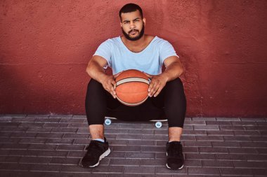 Koyu renkli sakallı adam portresi basketbol ile bir kaykay üzerinde oturan ve bir duvara yaslanmış bir beyaz gömlek ve spor şort giymiş.