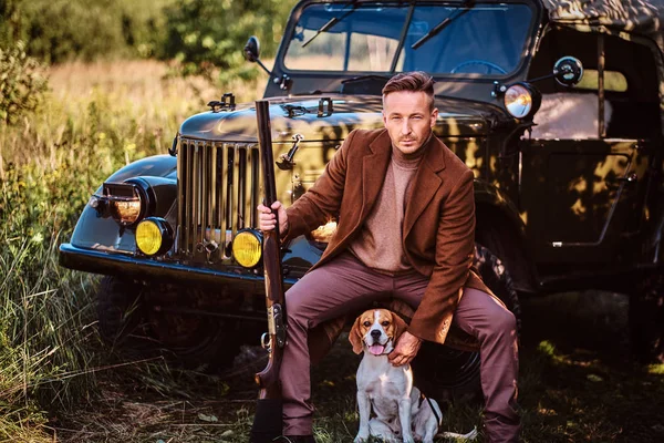 猎人穿着优雅的衣服, 手持猎枪, 坐在一起与他的猎犬狗, 而靠在一个复古军用车在森林. — 图库照片