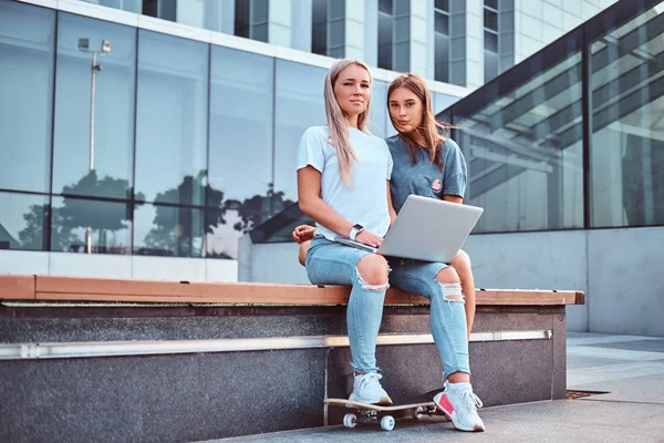 Dwie dziewczyny piękne hipster, siedząc na ławce z laptopa na tle drapacza chmur. — Zdjęcie stockowe