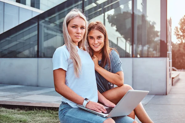 Két gyönyörű csípő lány ül a padon, egy laptop, a háttérben a felhőkarcoló. — Stock Fotó