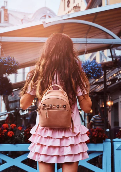 Вид сзади на женщину с каштановыми волосами, одетую в розовое платье с рюкзаком, стоящим рядом с кафе на террасе . — стоковое фото