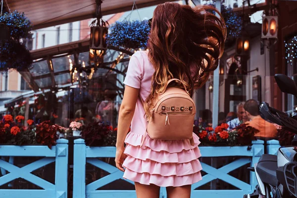 Rückansicht einer Frau mit braunen Haaren in rosa Kleid und einem Rucksack, die in der Nähe des Terrassencafés steht. — Stockfoto