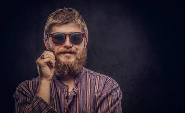 Happy hipster guy wearing sunglasses dressed in an old-fashioned shirt correct his mustache on a dark background. clipart