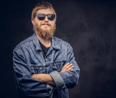 Güneş gözlüğü takıyor dalgın hipster adam koyu arka plan üzerinde çapraz silah ile poz kot ceket giymiş.