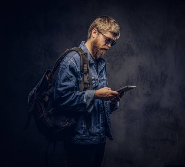 Güneş gözlüğü ve karanlık bir arka plan üzerinde bir tablet bilgisayar kullanarak kot ceket giymiş sırt çantası giyen hipster adam sakallı.