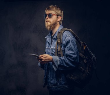 Sakallı hippi erkek güneş gözlüğü ve kot ceket giymiş sırt çantası takmış bir tablet bilgisayar karanlık bir arka plan üzerinde tutar..