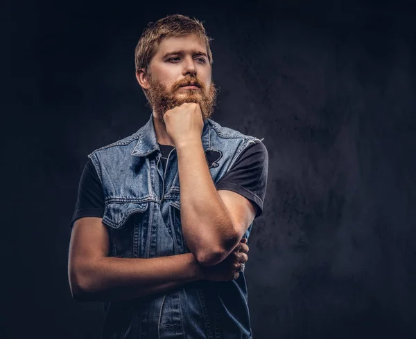 Retrato de un tipo hipster pensativo vestido con chaqueta vaquera posando con la mano en la barbilla sobre un fondo oscuro . — Foto de Stock