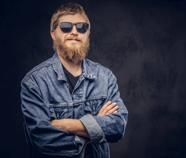 Nadenkend hipster vent dragen van een zonnebril gekleed in jeans jasje poseren met gekruiste armen op een donkere achtergrond. — Stockfoto