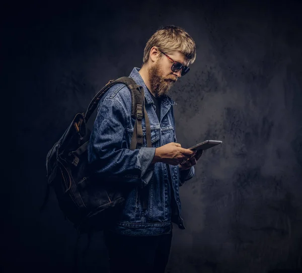 Tipo hipster barbudo con gafas de sol y mochila vestida con chaqueta vaquera usando una tableta sobre un fondo oscuro . —  Fotos de Stock