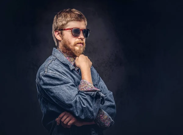 Pensive хіпстер хлопець в сонцезахисних окулярах, одягнений в джинсову куртку, позує рукою на підборідді на темному тлі . — стокове фото
