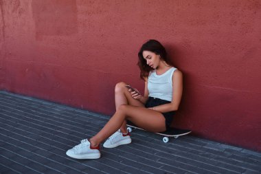 Bir smartphone kullanarak ve bir duvara yaslanmış iken müzik bir kaykay üzerinde oturan bir şehvetli hipster portresi.