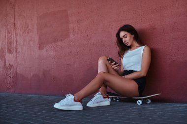 Bir smartphone kullanarak ve bir duvara yaslanmış iken müzik bir kaykay üzerinde oturan bir şehvetli hipster portresi.