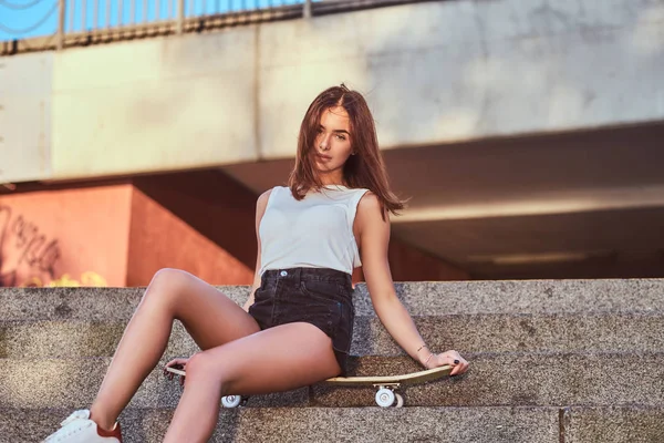 ショート パンツや橋の背景にステップでスケート ボードの上に座って シャツを着た美しい少女 — ストック写真