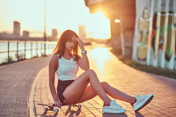 迷人的年轻时髦的女孩摆在坐在一个滑板在一个美丽的日落在路堤上 — 图库照片
