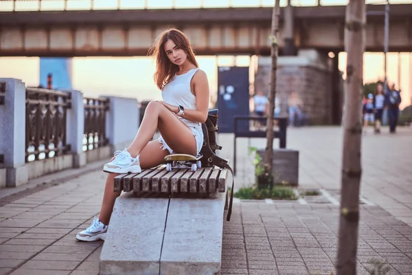 Jovem sensual skater menina vestida com shorts e t-shirt sentado no dique durante belo pôr do sol . — Fotografia de Stock