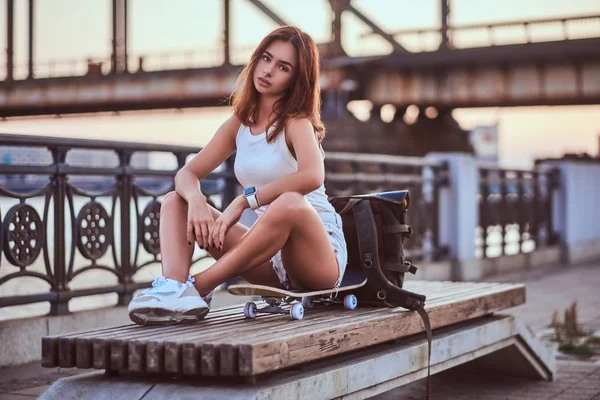 Joven chica patinadora sensual vestida con pantalones cortos y camiseta sentada en el terraplén durante el hermoso atardecer . — Foto de Stock